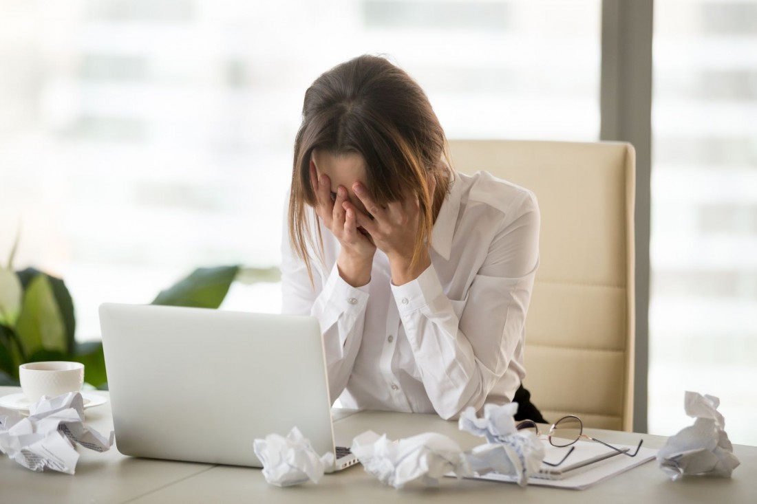 woman with her hands on her face in front of a laptop has several balls of paper on the table