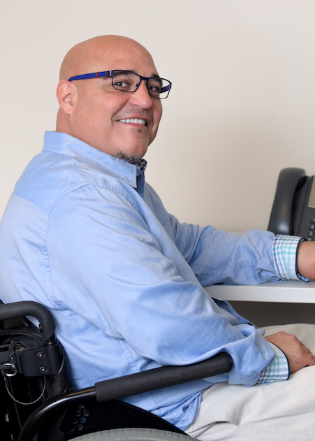 man in a wheelchair that is pointed away from camera, has his head turned to the camera and is smiling